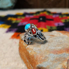 This eye-catching Navajo Turquoise & Coral Feather Ring features turquoise and red coral stones in sawtooth silver bezels with detailed feather, scroll, and silver bead accents; side view.