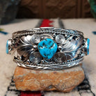 Navajo silver cuff featuring a central turquoise stone with dark matrix, surrounded by sculpted feather and leaf designs, silver spirals, twisted-wire borders, bead accents, and turquoise stones at the sides.