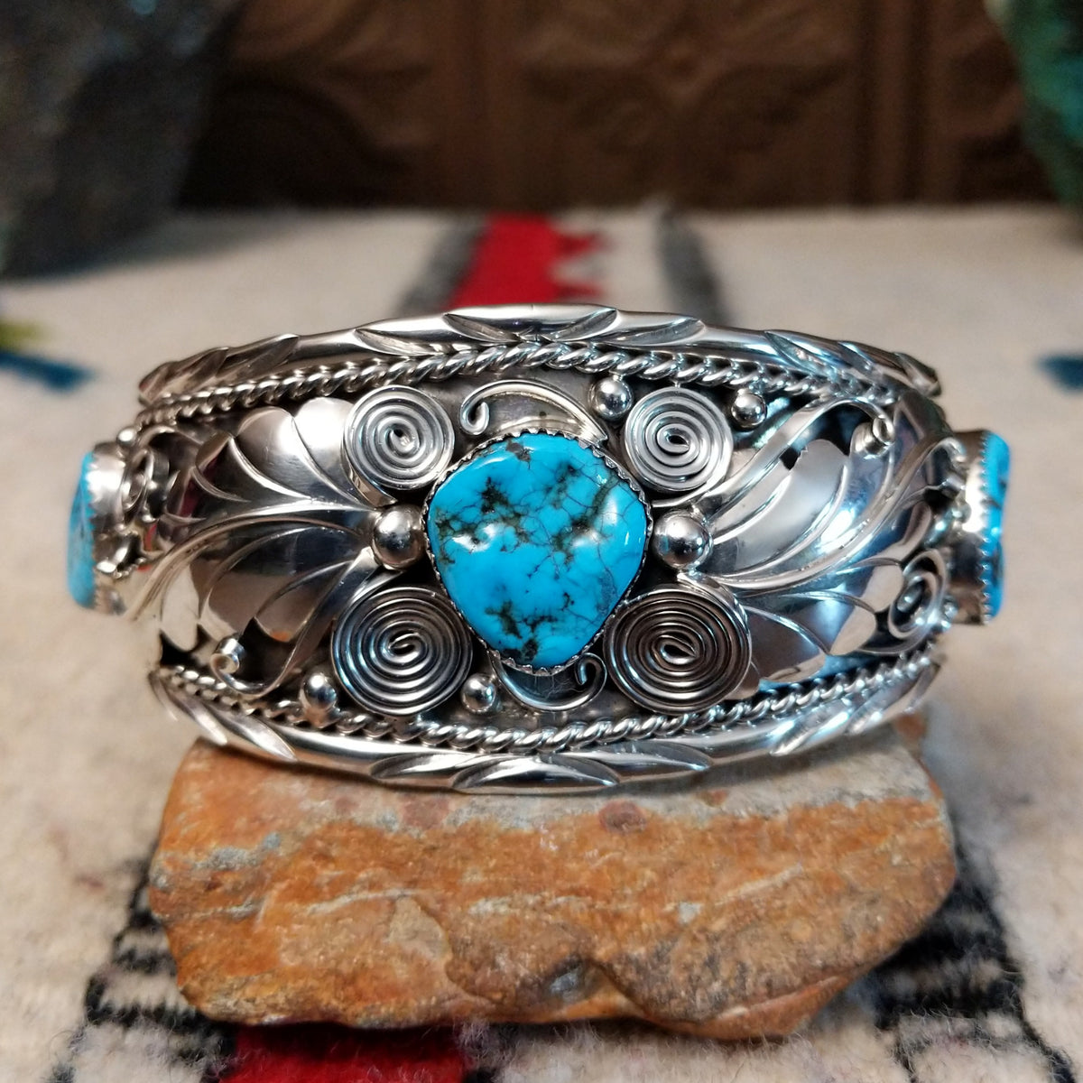 Navajo silver cuff featuring a central turquoise stone with dark matrix, surrounded by sculpted feather and leaf designs, silver spirals, twisted-wire borders, bead accents, and turquoise stones at the sides.