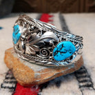 Navajo silver cuff featuring a central turquoise stone with dark matrix, surrounded by sculpted feather and leaf designs, silver spirals, twisted-wire borders, bead accents, and turquoise stones at the sides; side view.