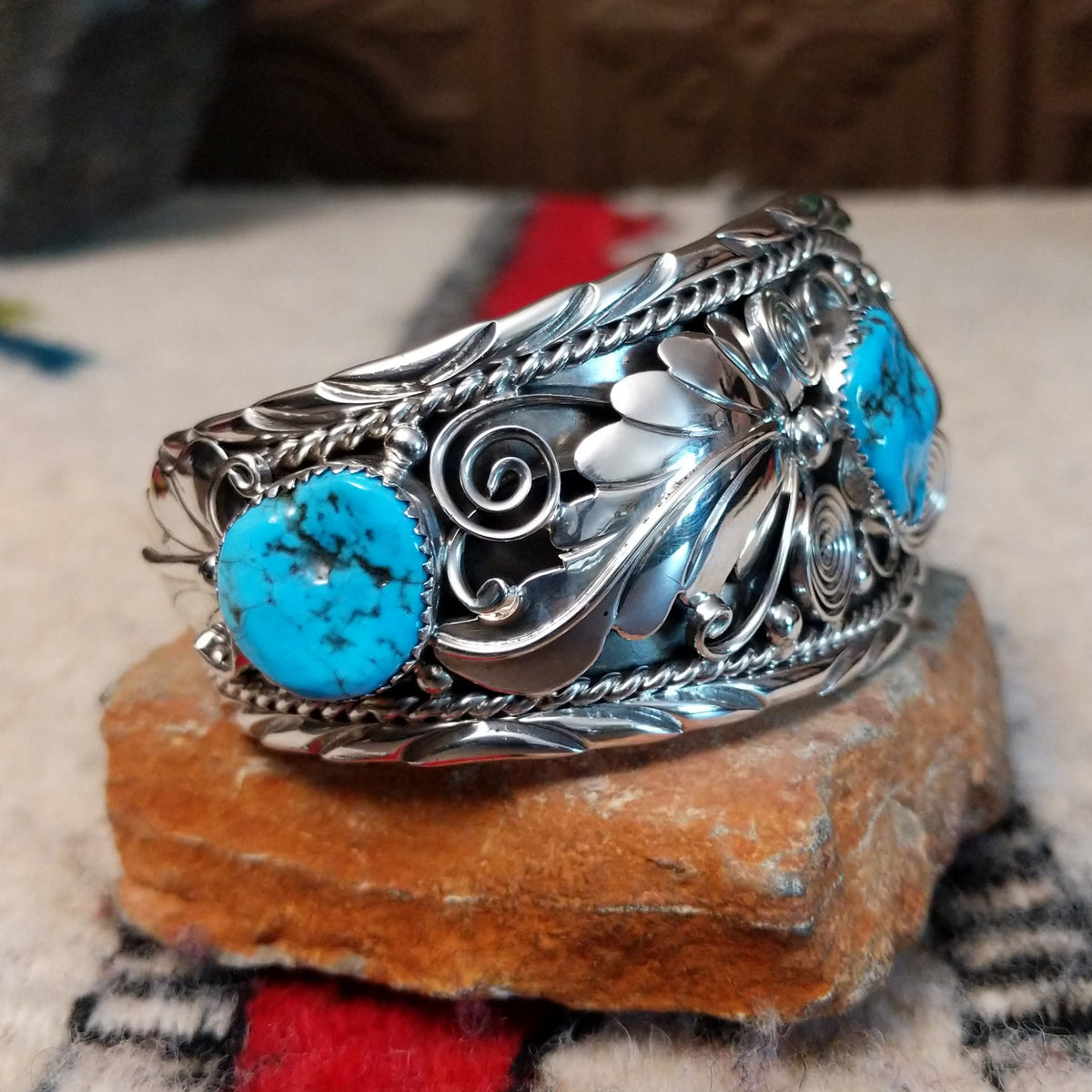 Navajo silver cuff featuring a central turquoise stone with dark matrix, surrounded by sculpted feather and leaf designs, silver spirals, twisted-wire borders, bead accents, and turquoise stones at the sides; side view.