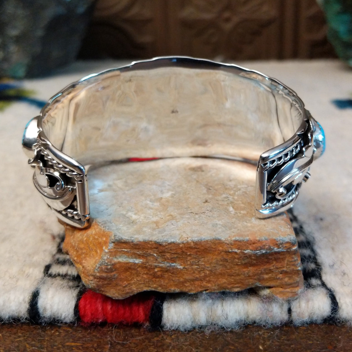 Navajo silver cuff featuring a central turquoise stone with dark matrix, surrounded by sculpted feather and leaf designs, silver spirals, twisted-wire borders, bead accents, and turquoise stones at the sides; underside view.
