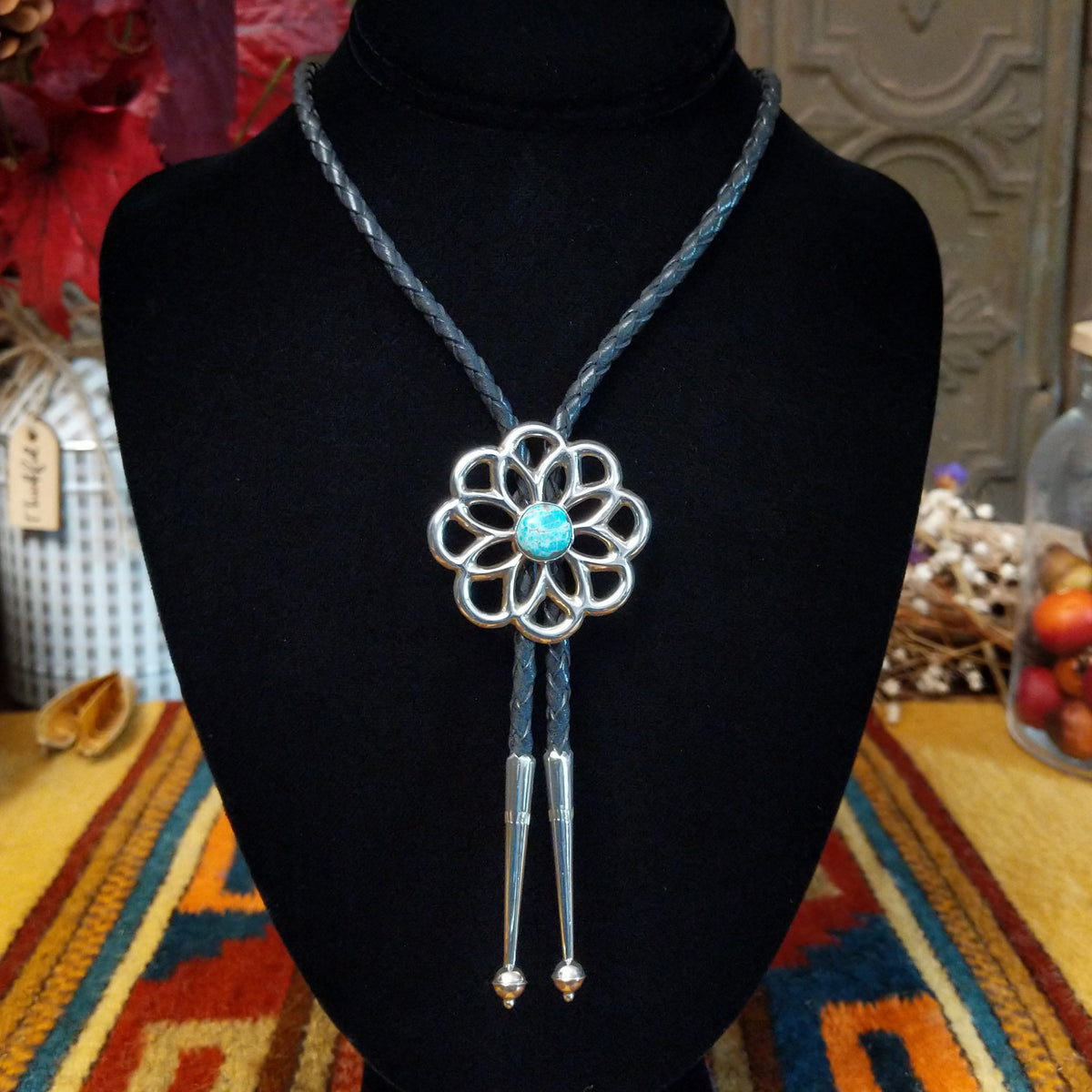 Navajo Turquoise Flower Bolo Tie by M. featuring an openwork silver flower with a round turquoise center stone, braided black leather cord, and polished silver tips.