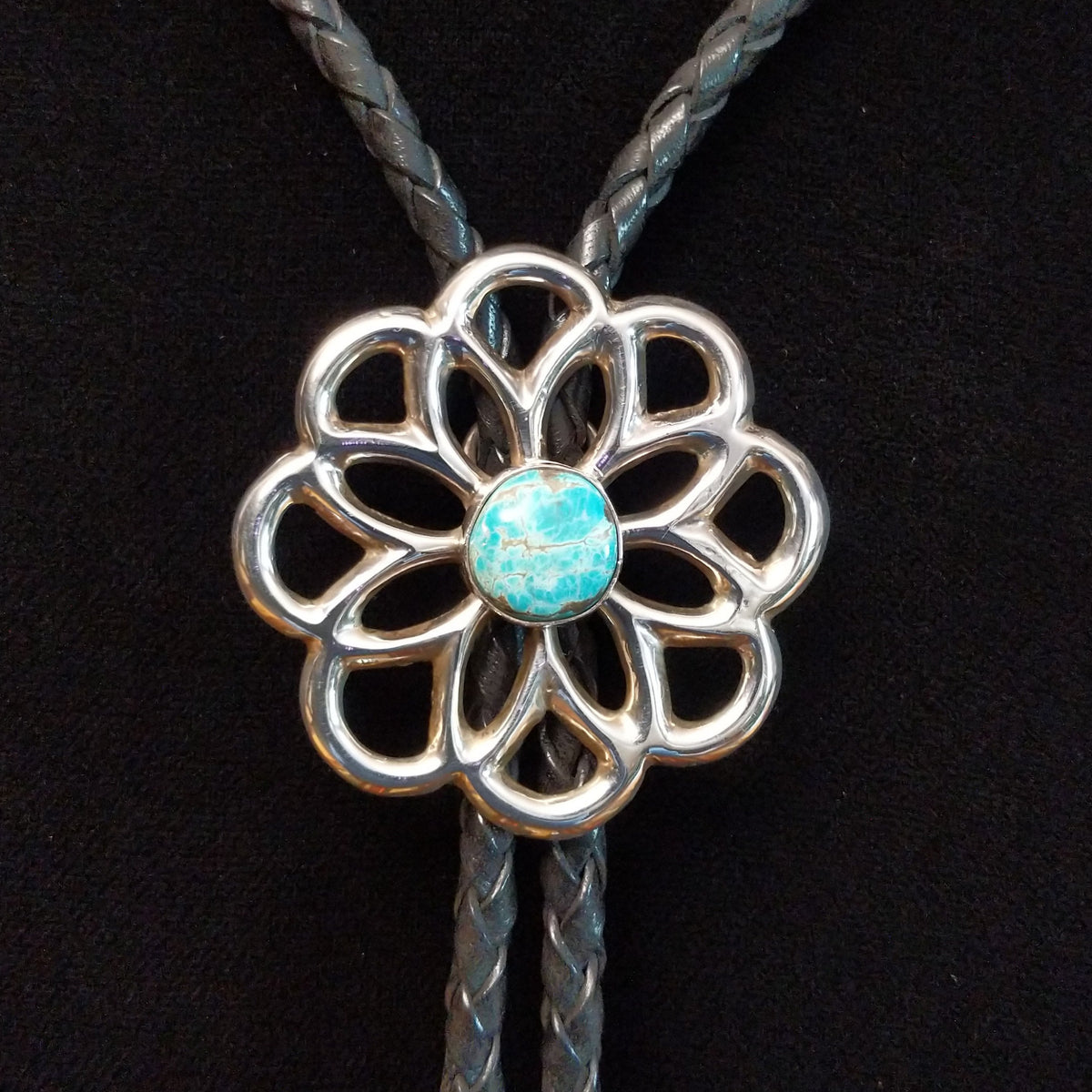 Navajo Turquoise Flower Bolo Tie by M. featuring an openwork silver flower with a round turquoise center stone, braided black leather cord, and polished silver tips.