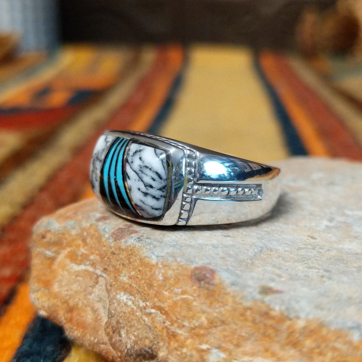 This elegant Navajo White Buffalo with Turquoise Inlay Ring features a rectangular silver setting with black-and-white White Buffalo stone and vibrant turquoise inlay arranged in a flowing geometric pattern; side view.