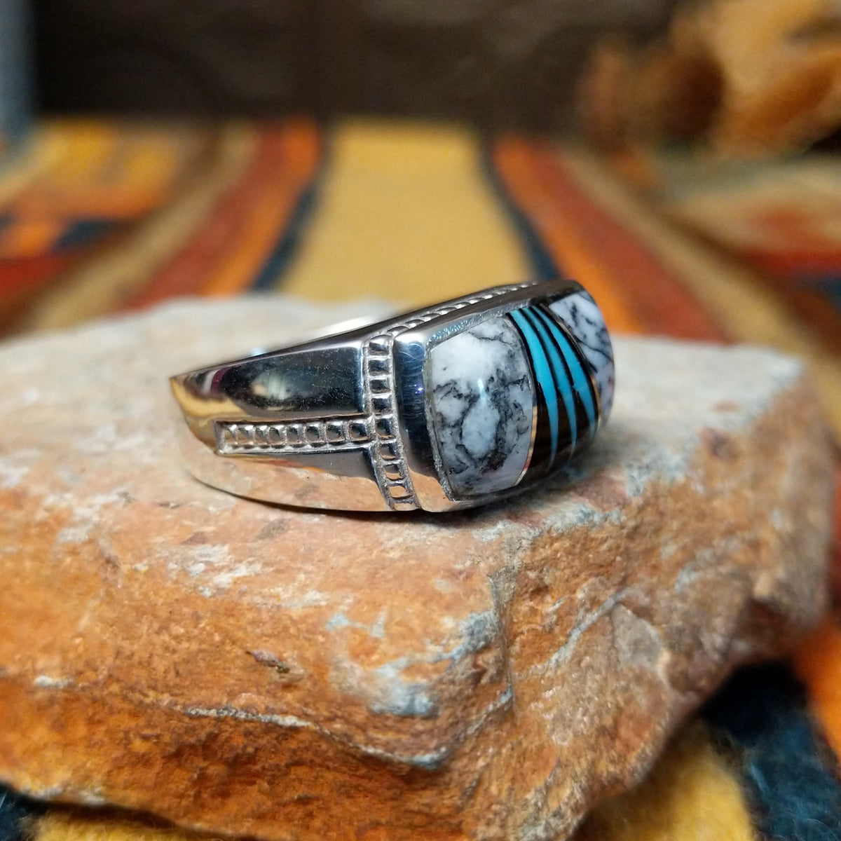 This elegant Navajo White Buffalo with Turquoise Inlay Ring features a rectangular silver setting with black-and-white White Buffalo stone and vibrant turquoise inlay arranged in a flowing geometric pattern; side view.