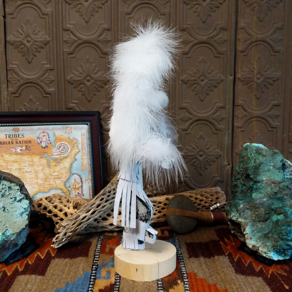 Wood carved into a cloud kachina in vibrant white paint with black details along the body, a feather headdress, and is set on a wooden base.