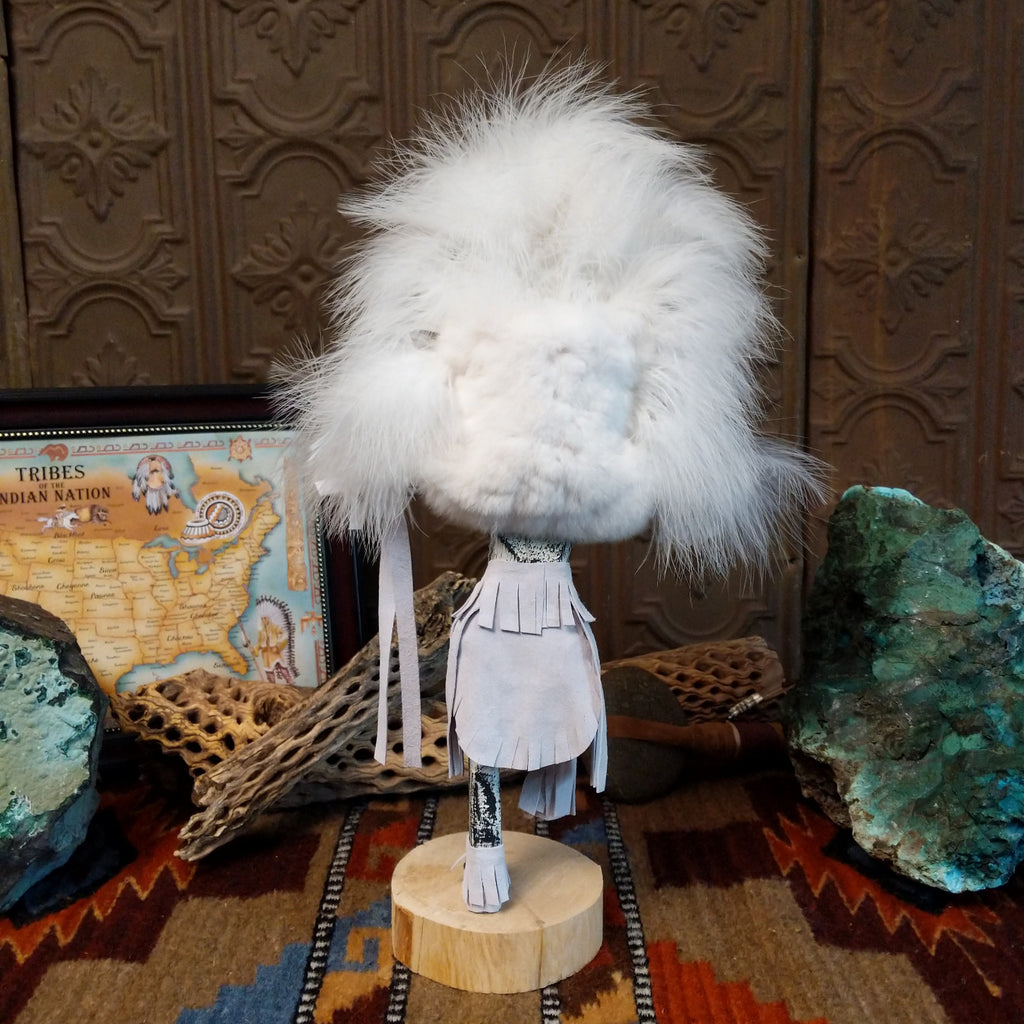 Wood carved into a cloud kachina in vibrant white paint with black details along the body, a feather headdress, and is set on a wooden base.