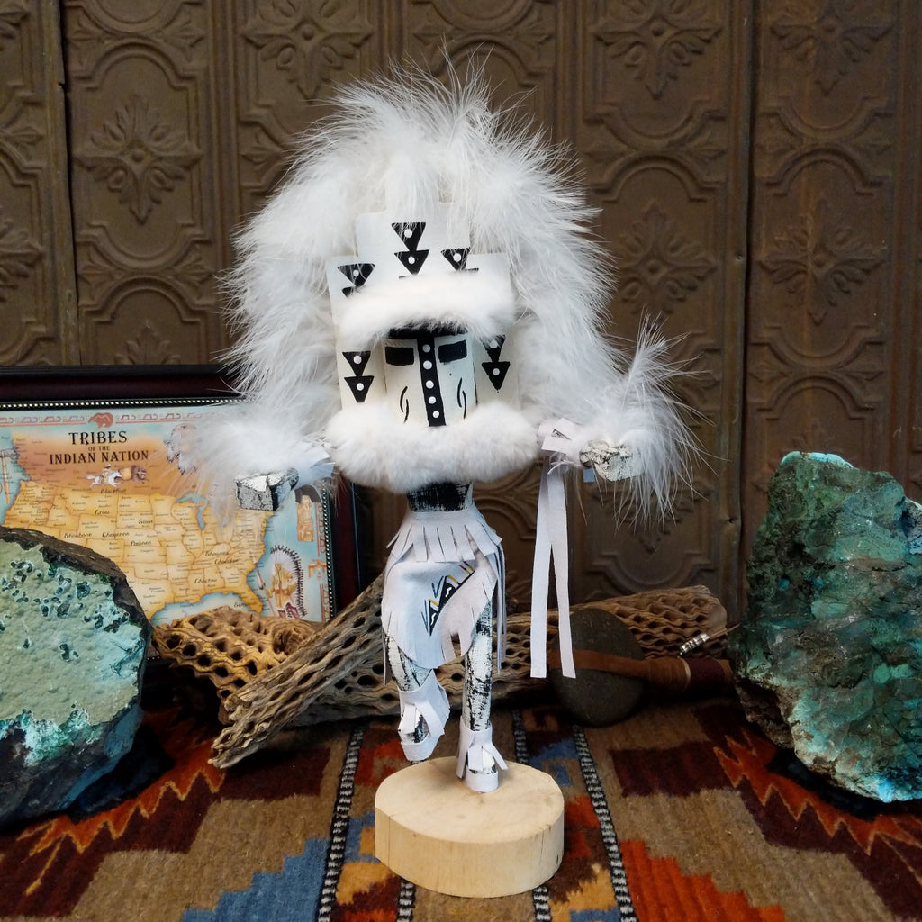 Wood carved into a cloud kachina in vibrant white paint with black details along the body, a feather headdress, and is set on a wooden base.
