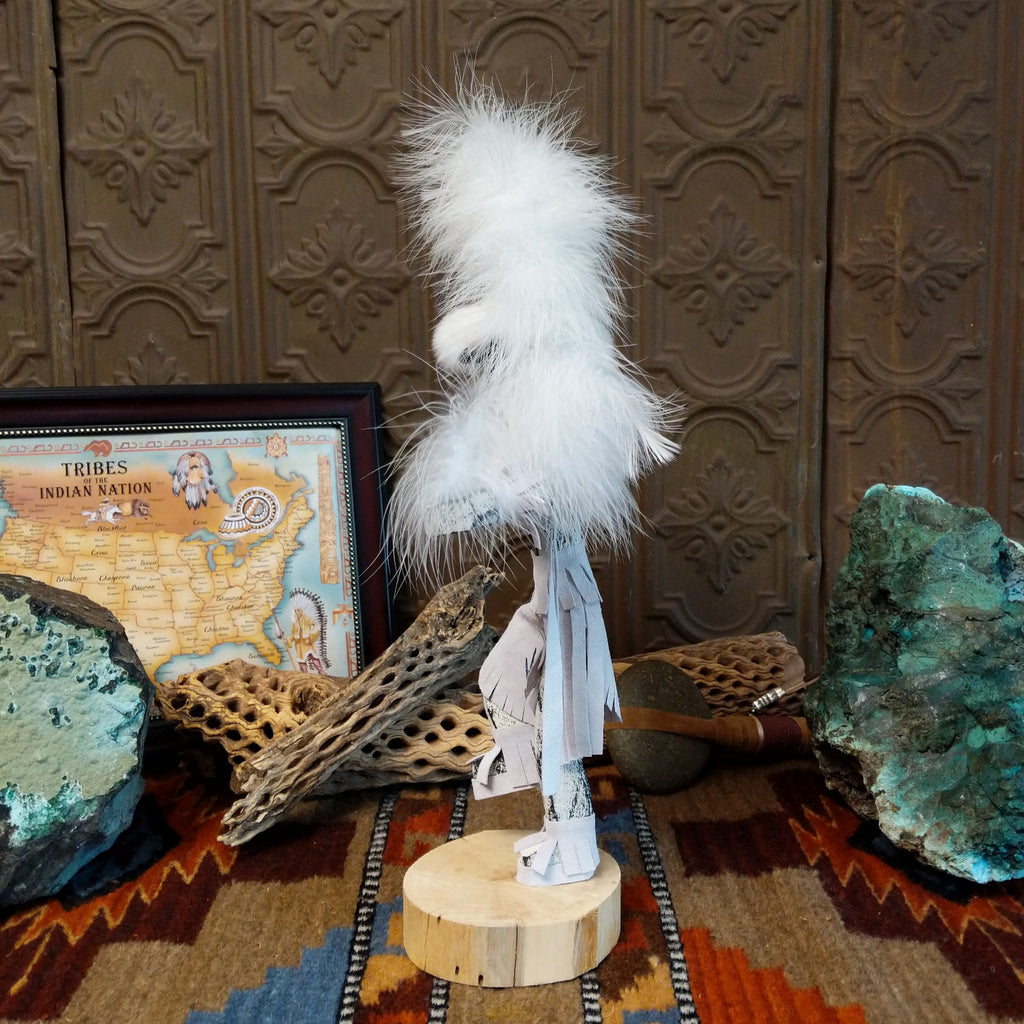 Wood carved into a cloud kachina in vibrant white paint with black details along the body, a feather headdress, and is set on a wooden base.