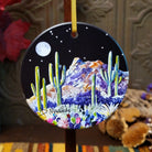 A round hanging ornament with a hand-painted desert night scene, featuring saguaro cacti, purple flowers, mountains, a moon, and stars on a black background. The ornament is suspended by a yellow ribbon, with a blurred rustic background.