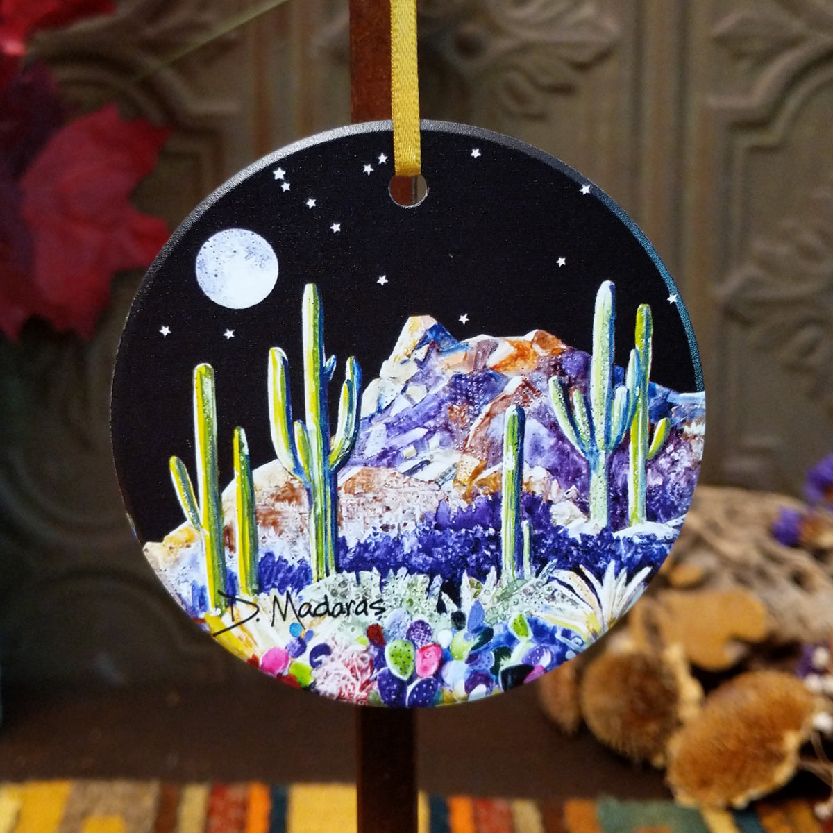 A round hanging ornament with a hand-painted desert night scene, featuring saguaro cacti, purple flowers, mountains, a moon, and stars on a black background. The ornament is suspended by a yellow ribbon, with a blurred rustic background.