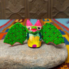 This colorful Oaxacan bat wood carving features green patterned wings, bright pink ears, expressive eyes, and a hand-painted yellow floral design.