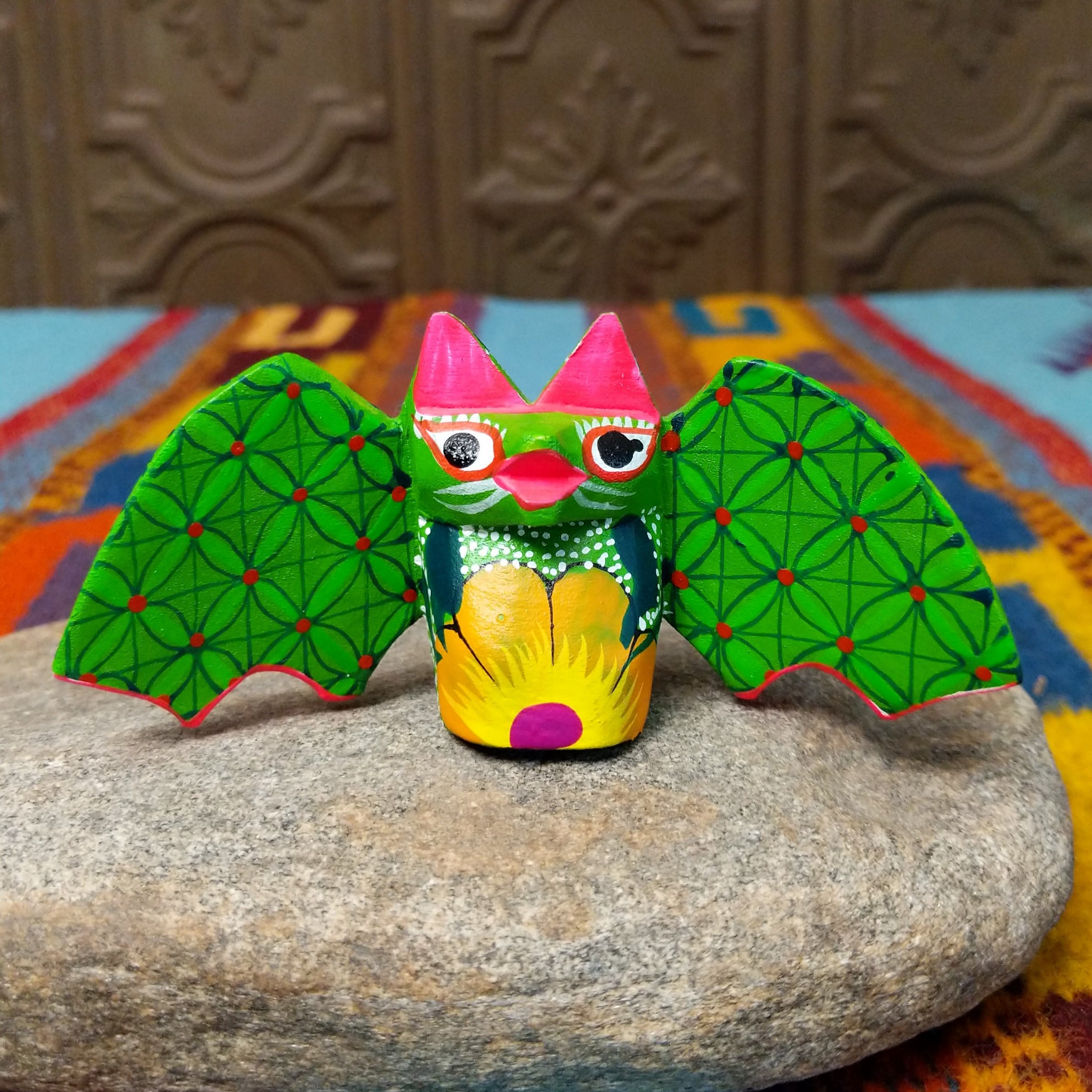 This colorful Oaxacan bat wood carving features green patterned wings, bright pink ears, expressive eyes, and a hand-painted yellow floral design.