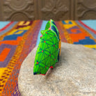 This colorful Oaxacan bat wood carving features green patterned wings, bright pink ears, expressive eyes, and a hand-painted yellow floral design; side view.