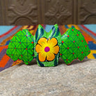 This colorful Oaxacan bat wood carving features green patterned wings, bright pink ears, expressive eyes, and a hand-painted yellow floral design; rear view.