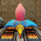 Oaxacan bird wood carving with a vibrant yellow head, orange beak, and expansive blue wings adorned with intricate patterns.
