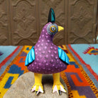 Oaxacan bird wood carving by Hedilberto Olivera with a purple body, broad blue wings, fan-shaped crest, and yellow beak and feet.