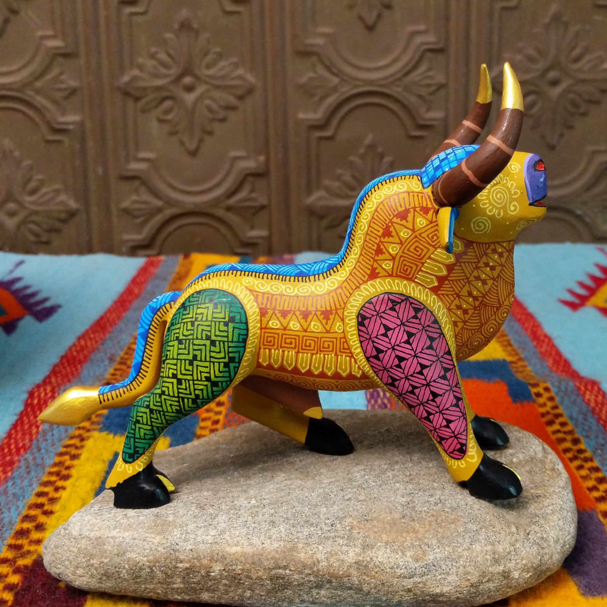 Oaxacan bull wood carving by Abundio Muñoz with curved horns, a golden yellow body, and colorful geometric patterns across the legs and torso.