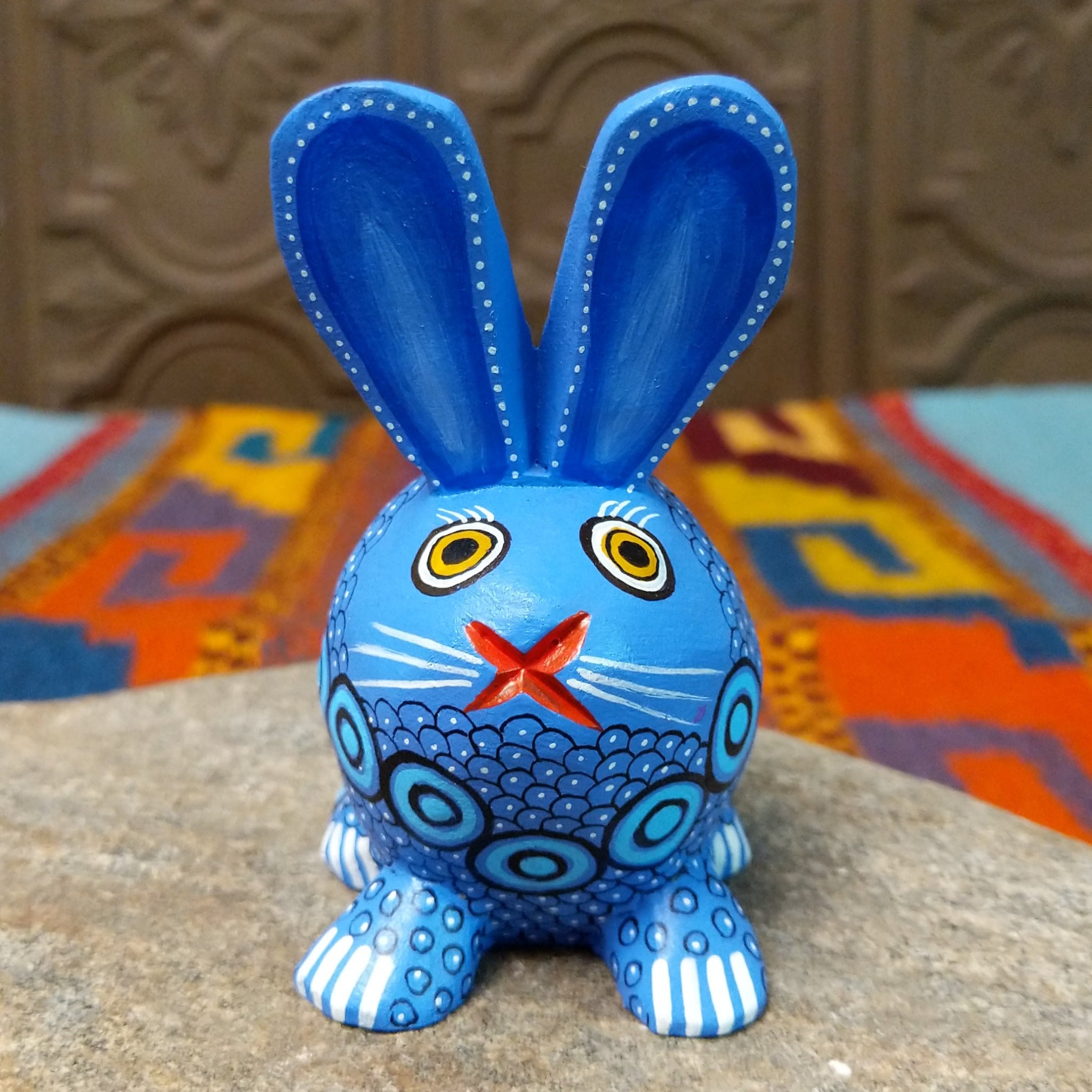 Blue Oaxacan bunny wood carving by Alberto Hernandez with tall ears, ringed eyes, and layered circular and scale-like painted patterns.