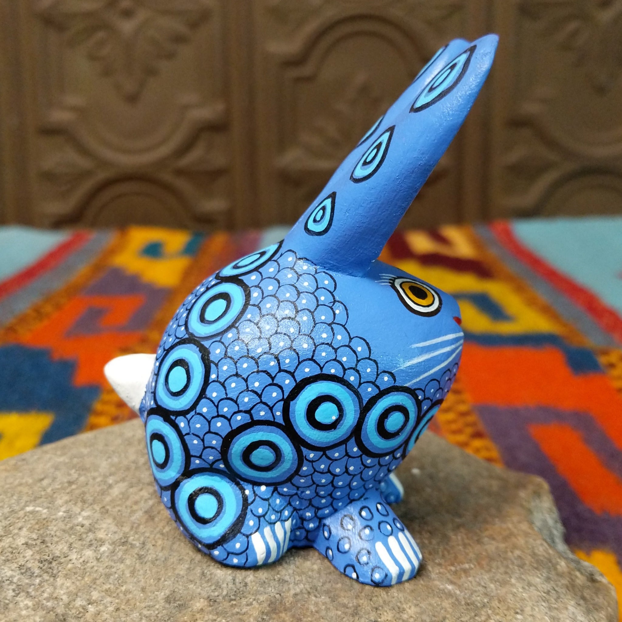 Blue Oaxacan bunny wood carving by Alberto Hernandez with tall ears, ringed eyes, and layered circular and scale-like painted patterns.