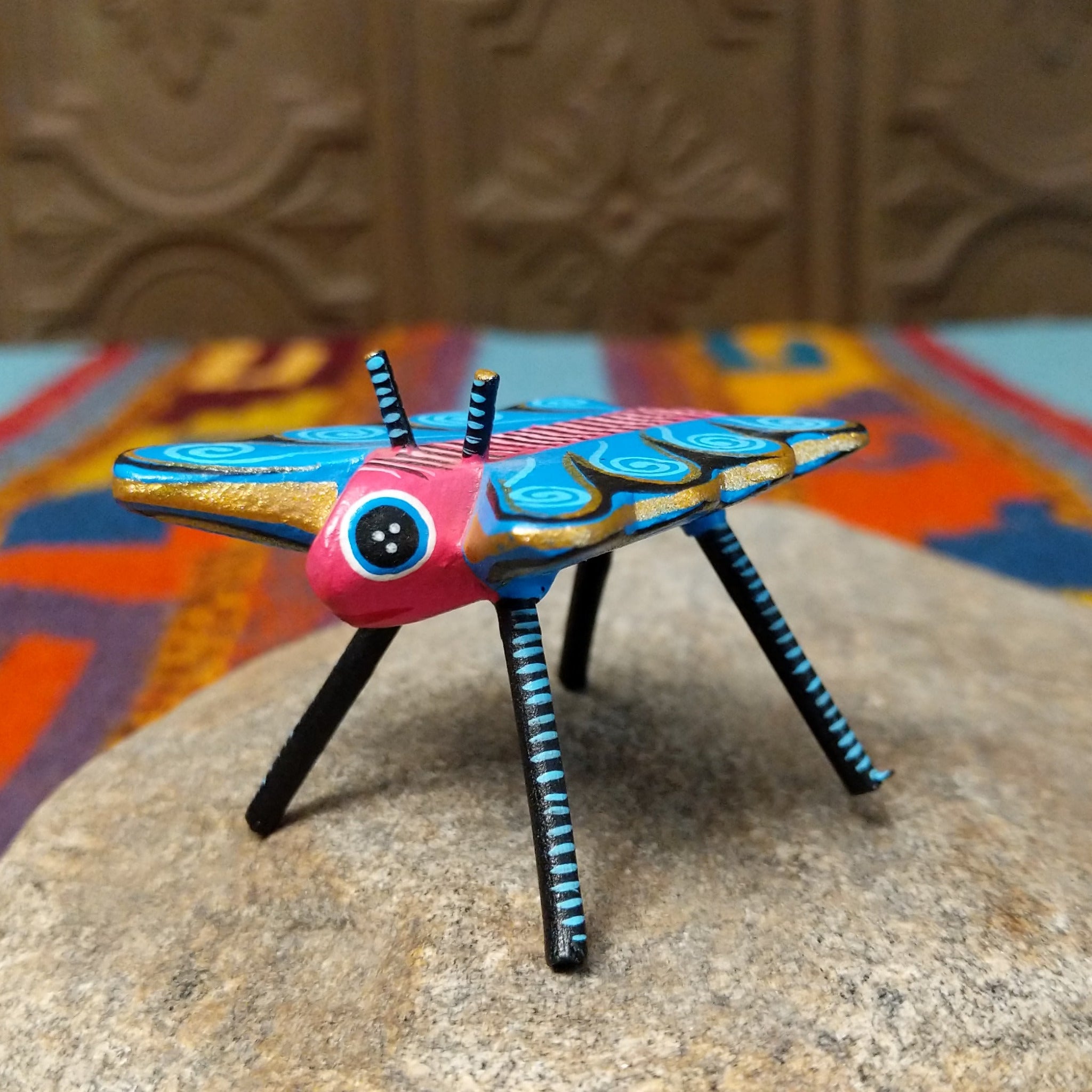 This colorful Oaxacan butterfly wood carving features a pink body, turquoise wings with spiral patterns, gold accents, striped antennae, and long black-and-turquoise legs.