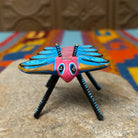 This colorful Oaxacan butterfly wood carving features a pink body, turquoise wings with spiral patterns, gold accents, striped antennae, and long black-and-turquoise legs; front facing view.
