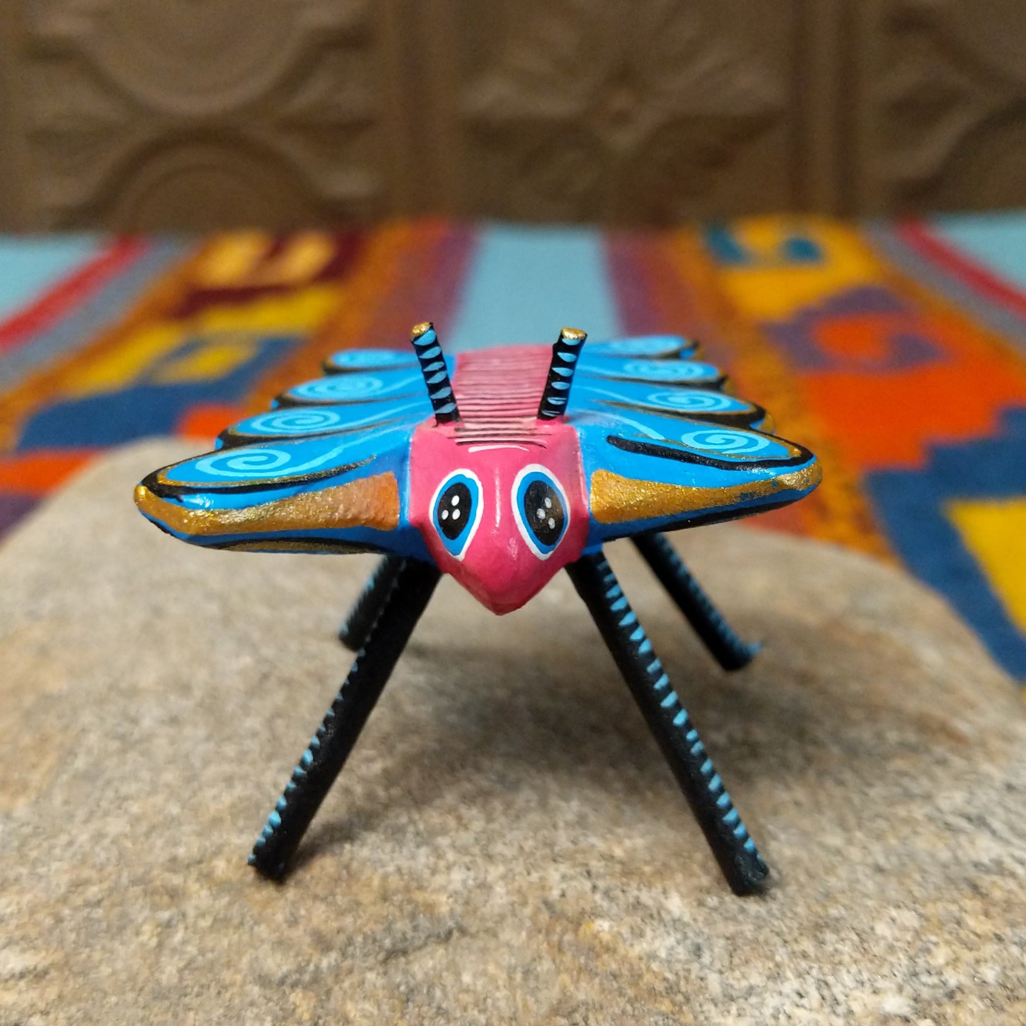This colorful Oaxacan butterfly wood carving features a pink body, turquoise wings with spiral patterns, gold accents, striped antennae, and long black-and-turquoise legs; front facing view.