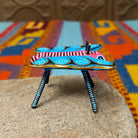 This colorful Oaxacan butterfly wood carving features a pink body, turquoise wings with spiral patterns, gold accents, striped antennae, and long black-and-turquoise legs; side view.
