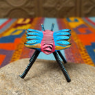 This colorful Oaxacan butterfly wood carving features a pink body, turquoise wings with spiral patterns, gold accents, striped antennae, and long black-and-turquoise legs; rear view.