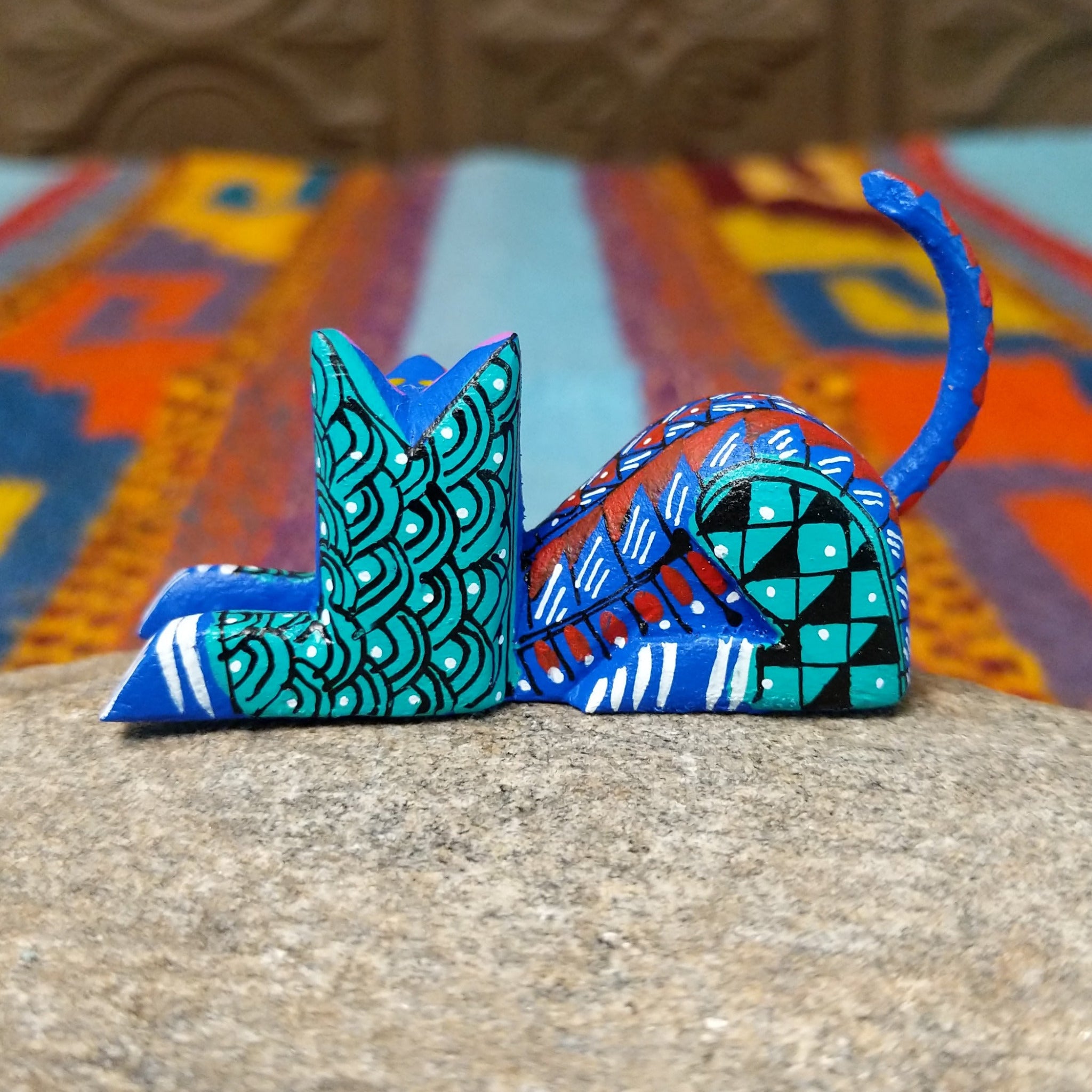 Reclining Oaxacan cat wood carving with a curled tail, oversized eyes, and colorful hand-painted geometric patterns.