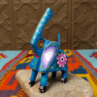 This blue Oaxacan elephant wood carving by Roberta Angeles features an uplifted trunk, lavender ear, pink flower, and colorful hand-painted decorative patterns.