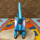 This blue Oaxacan elephant wood carving by Roberta Angeles features an uplifted trunk, lavender ear, pink flower, and colorful hand-painted decorative patterns; front view.