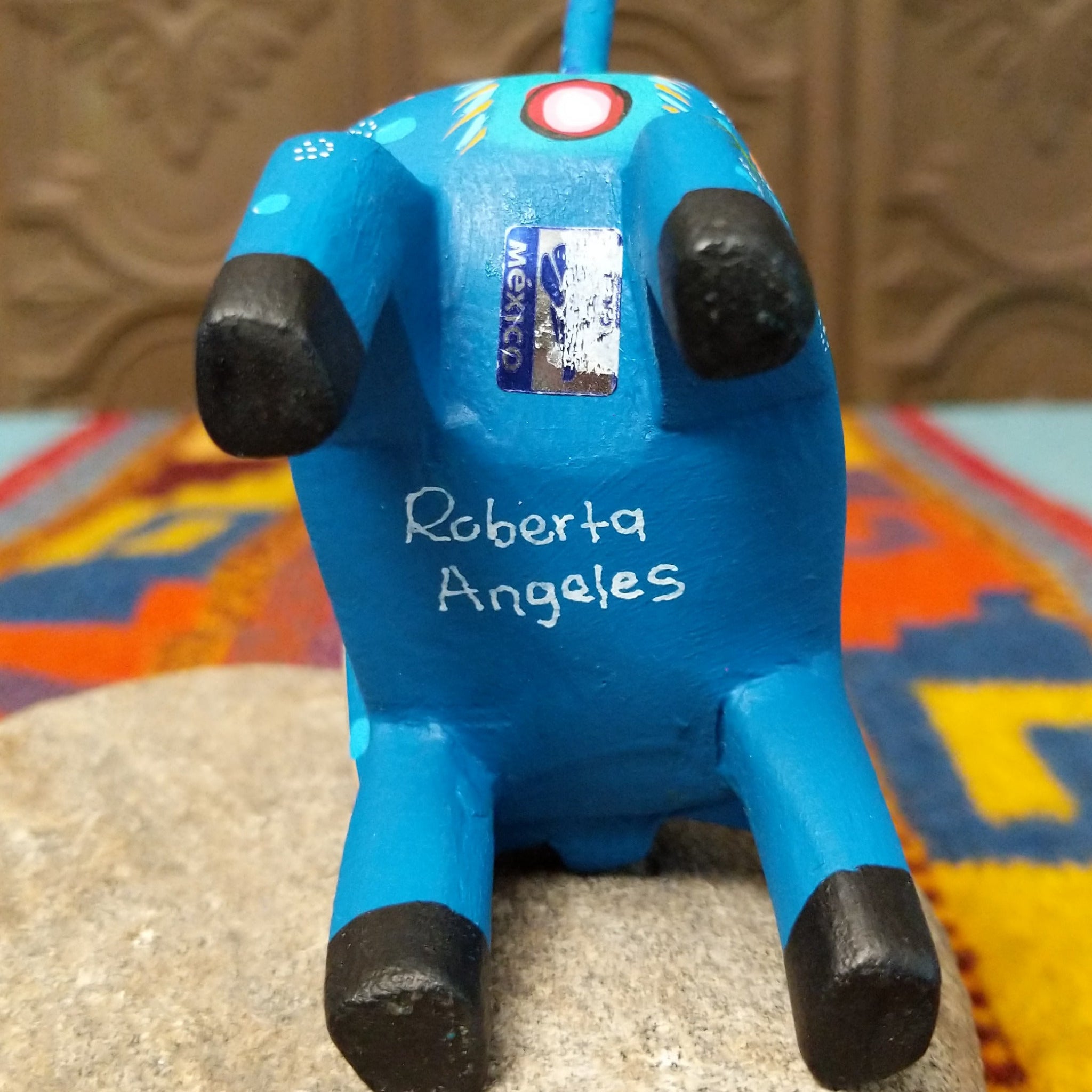 This blue Oaxacan elephant wood carving by Roberta Angeles features an uplifted trunk, lavender ear, pink flower, and colorful hand-painted decorative patterns; artisan name and made in Mexico sticker.