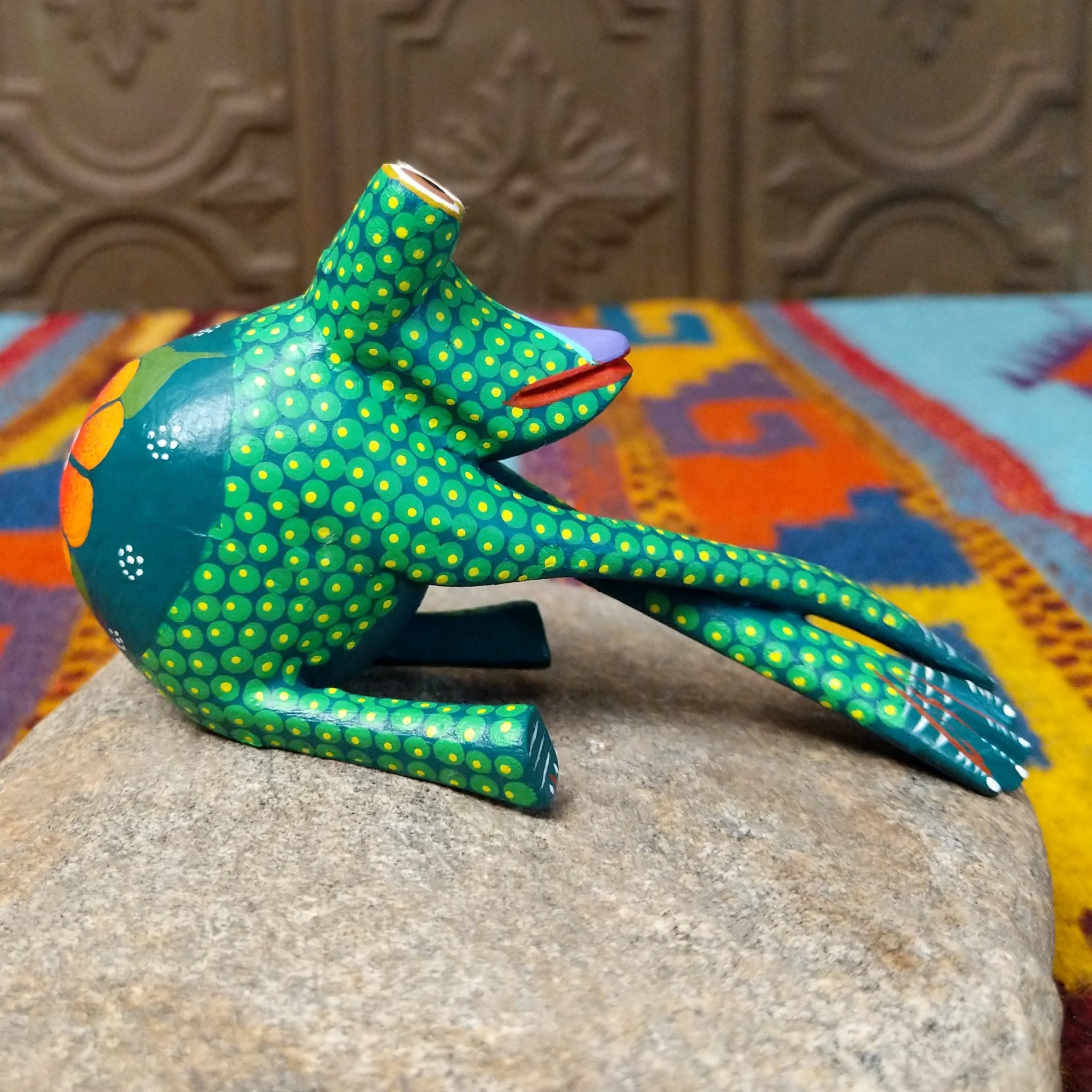 This green Oaxacan frog wood carving by Roberta Angeles features raised eyes, colorful dotted patterns, a red mouth, and long legs with oversized hand-painted webbed feet; side view.