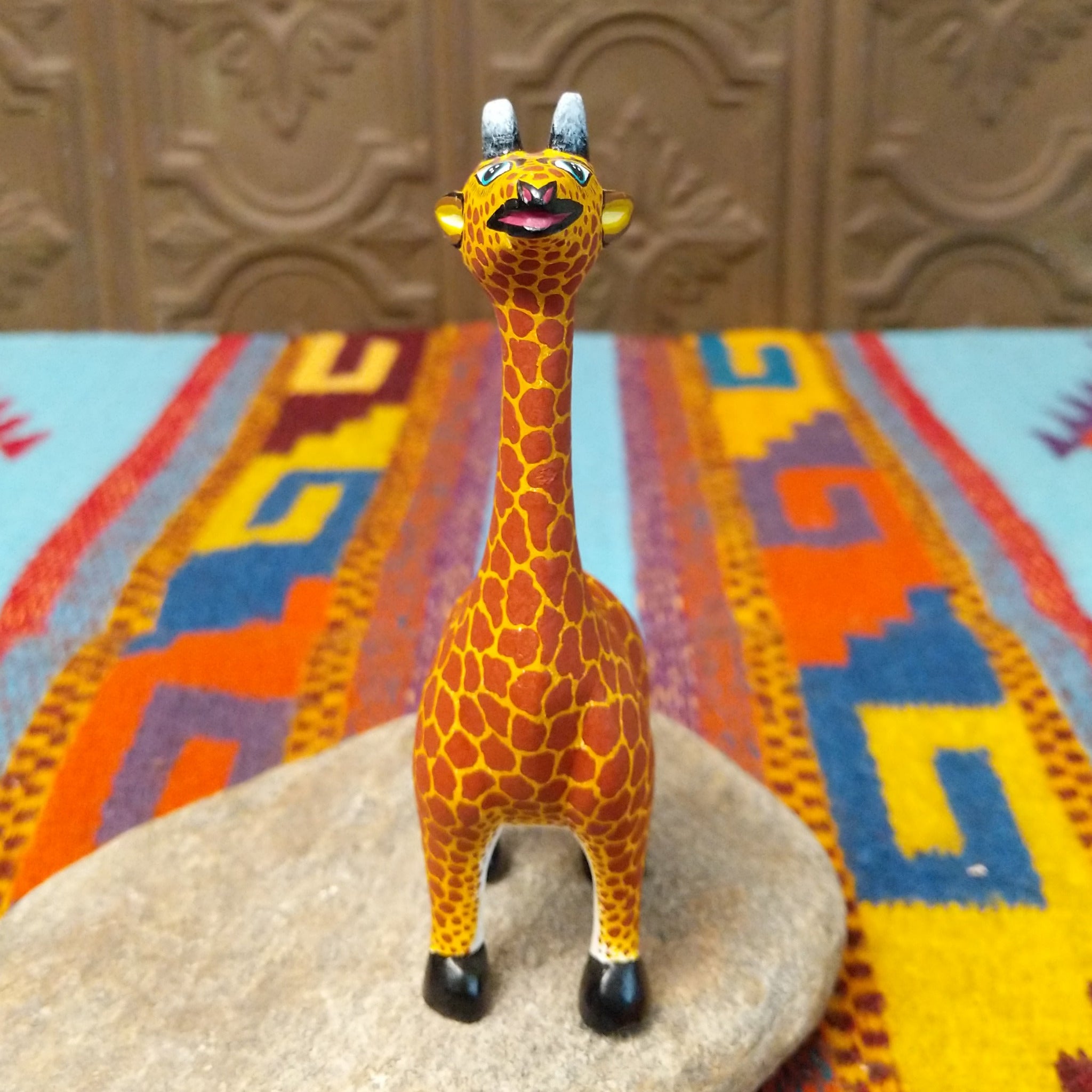 This Oaxacan giraffe wood carving by Faviola Fabian features a golden-yellow body with reddish-brown spots, a long neck, blue eyes, black-and-white horns, and black hooves; front facing view.
