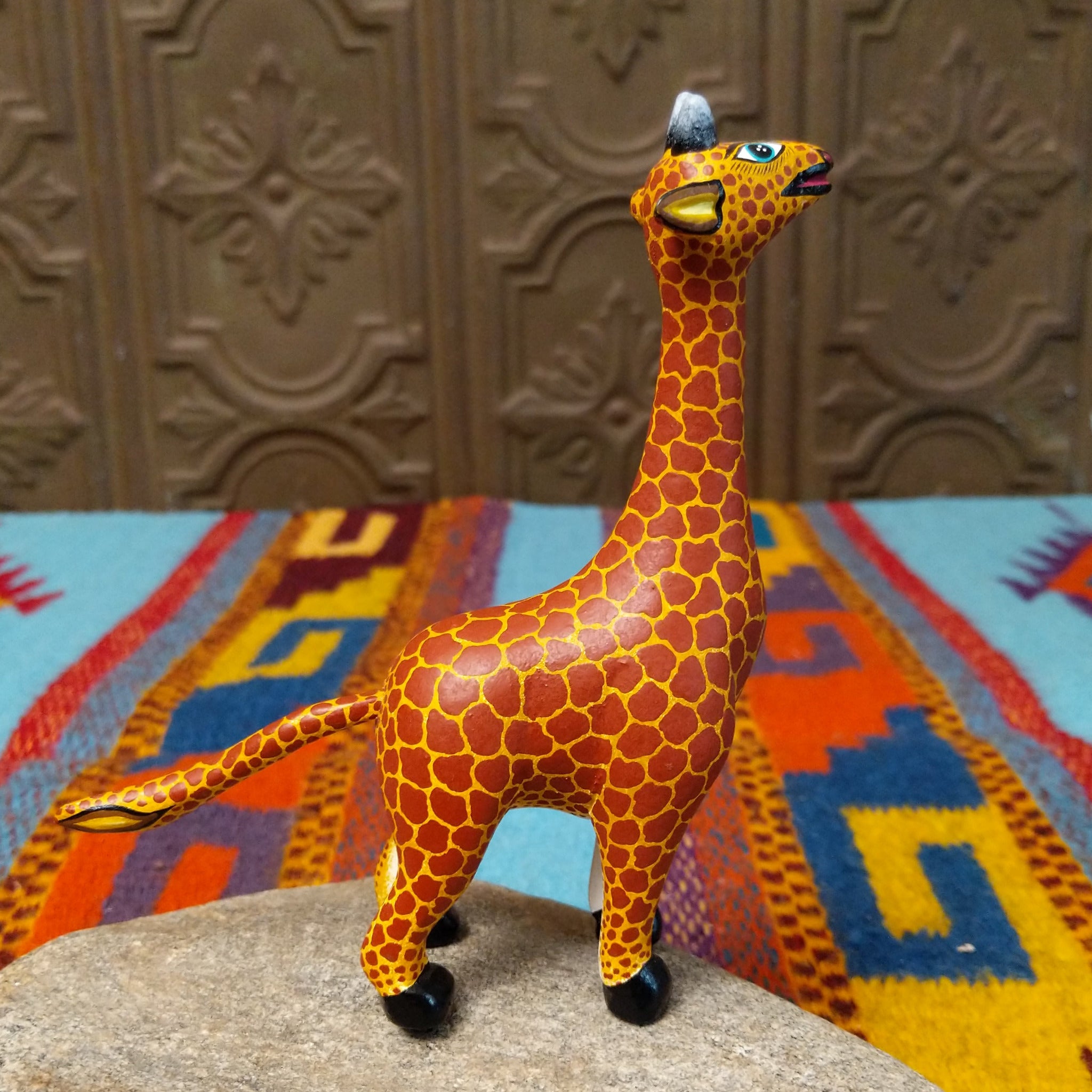 This Oaxacan giraffe wood carving by Faviola Fabian features a golden-yellow body with reddish-brown spots, a long neck, blue eyes, black-and-white horns, and black hooves; side view.