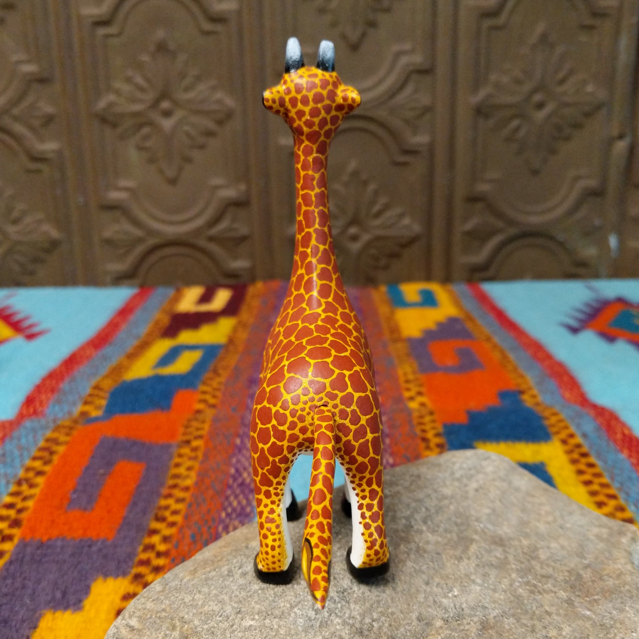 This Oaxacan giraffe wood carving by Faviola Fabian features a golden-yellow body with reddish-brown spots, a long neck, blue eyes, black-and-white horns, and black hooves; rear view.