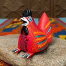 This colorful Oaxacan hen wood carving features a bright red body, black comb, turquoise eye, and intricate yellow, orange, turquoise, and black hand-painted feather patterns.