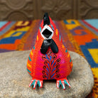 This colorful Oaxacan hen wood carving features a bright red body, black comb, turquoise eye, and intricate yellow, orange, turquoise, and black hand-painted feather patterns; front facing view.