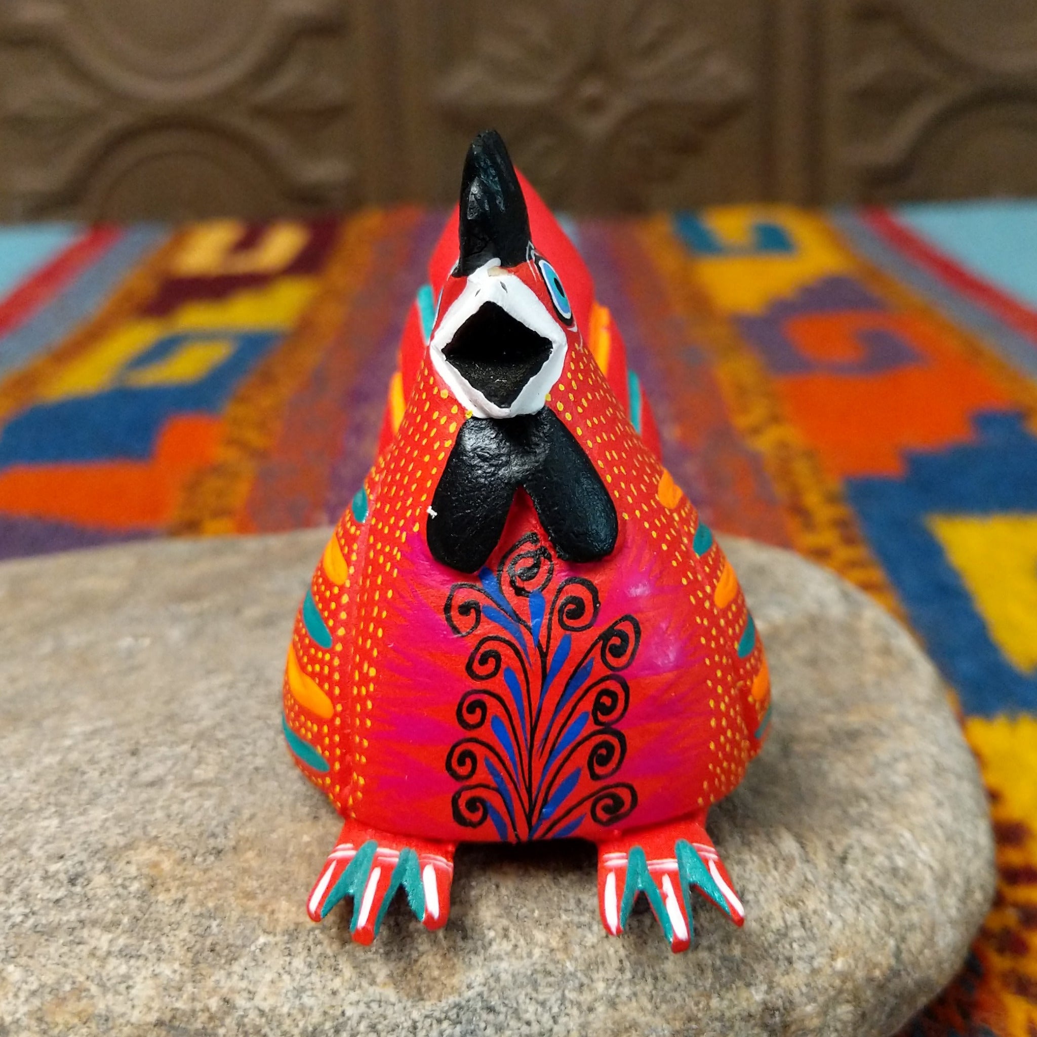 This colorful Oaxacan hen wood carving features a bright red body, black comb, turquoise eye, and intricate yellow, orange, turquoise, and black hand-painted feather patterns; front facing view.