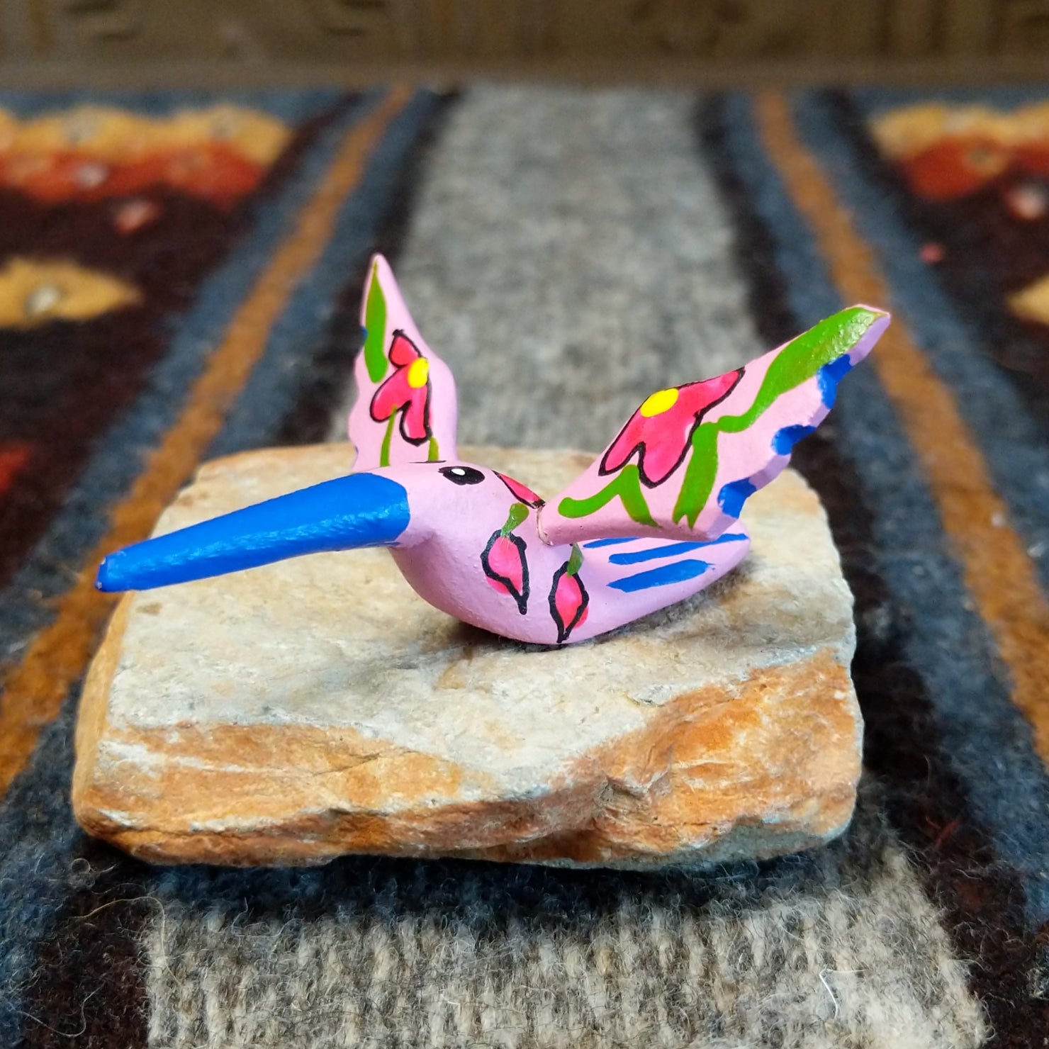 Oaxacan hummingbird carving painted in soft pink, with a striking blue beak and red floral accents on its wings.