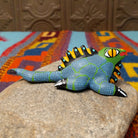 This colorful Oaxacan iguana wood carving features a green head, blue dotted body, golden eyes, black-and-yellow back spines, and a long patterned tail; side view.