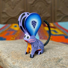 Oaxacan jerboa wood carving with oversized blue patterned ears, a lavender body, orange front legs, and long curved black tail.