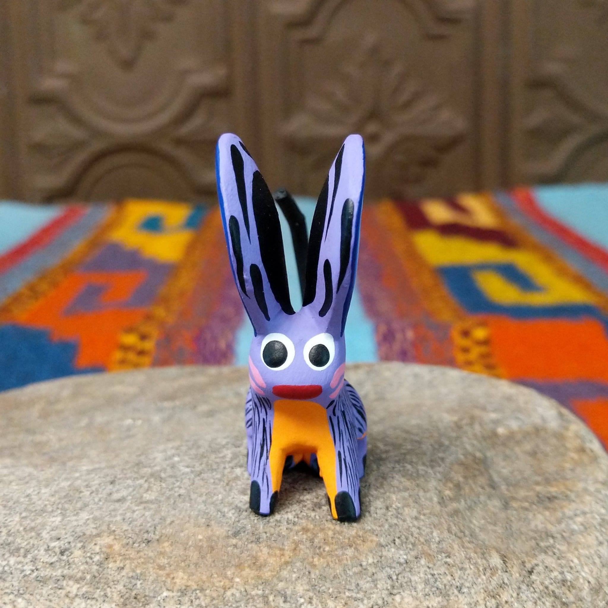 Oaxacan jerboa wood carving with oversized blue patterned ears, a lavender body, orange front legs, and long curved black tail.