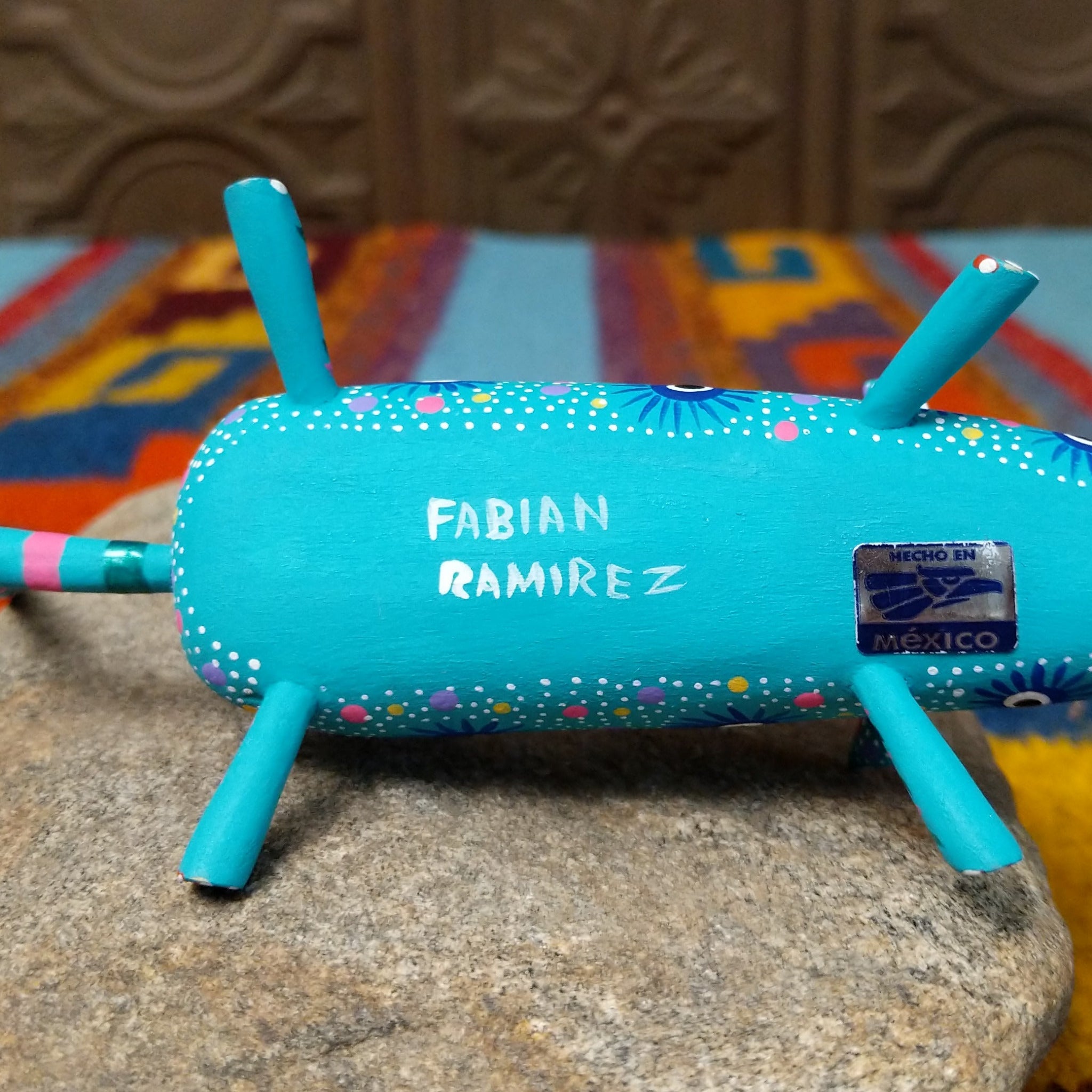 This turquoise Oaxacan mouse wood carving by Fabian Ramirez features colorful hand-painted patterns, pink-accented ears and legs, and a long striped tail; artisan name and made in Mexico sticker.