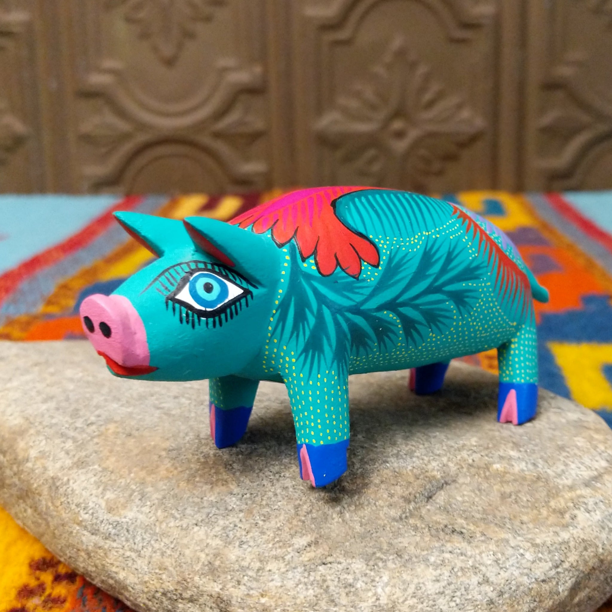 This turquoise Oaxacan pig wood carving features a pink snout, expressive blue eyes, colorful floral and leaf patterns, and blue-and-pink painted hooves.