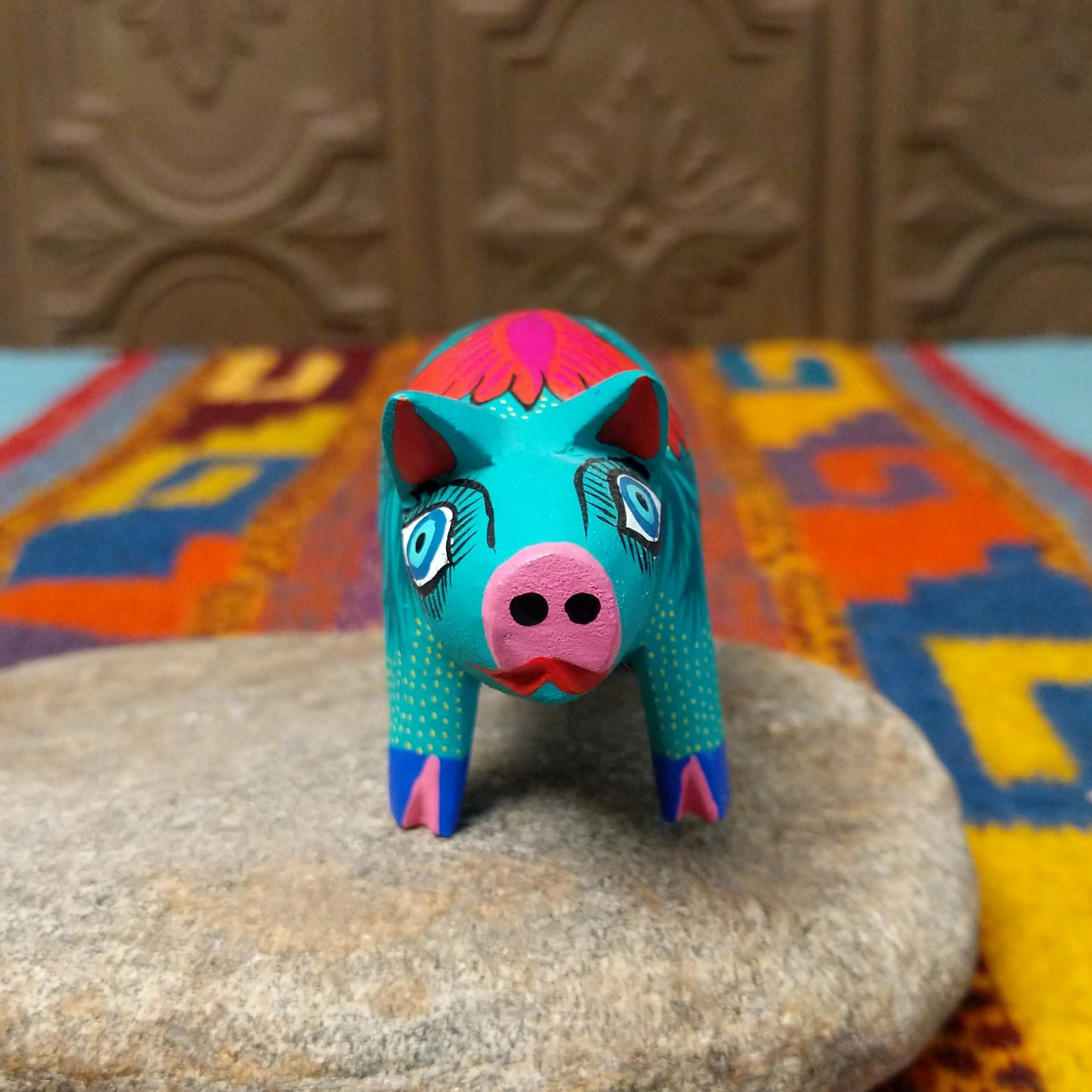 This turquoise Oaxacan pig wood carving features a pink snout, expressive blue eyes, colorful floral and leaf patterns, and blue-and-pink painted hooves; front view.