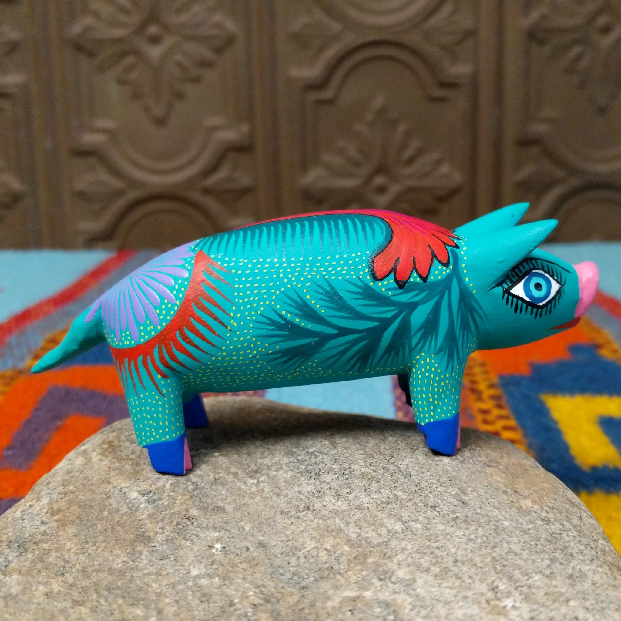 This turquoise Oaxacan pig wood carving features a pink snout, expressive blue eyes, colorful floral and leaf patterns, and blue-and-pink painted hooves; side view.