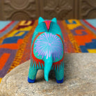 This turquoise Oaxacan pig wood carving features a pink snout, expressive blue eyes, colorful floral and leaf patterns, and blue-and-pink painted hooves; rear view.